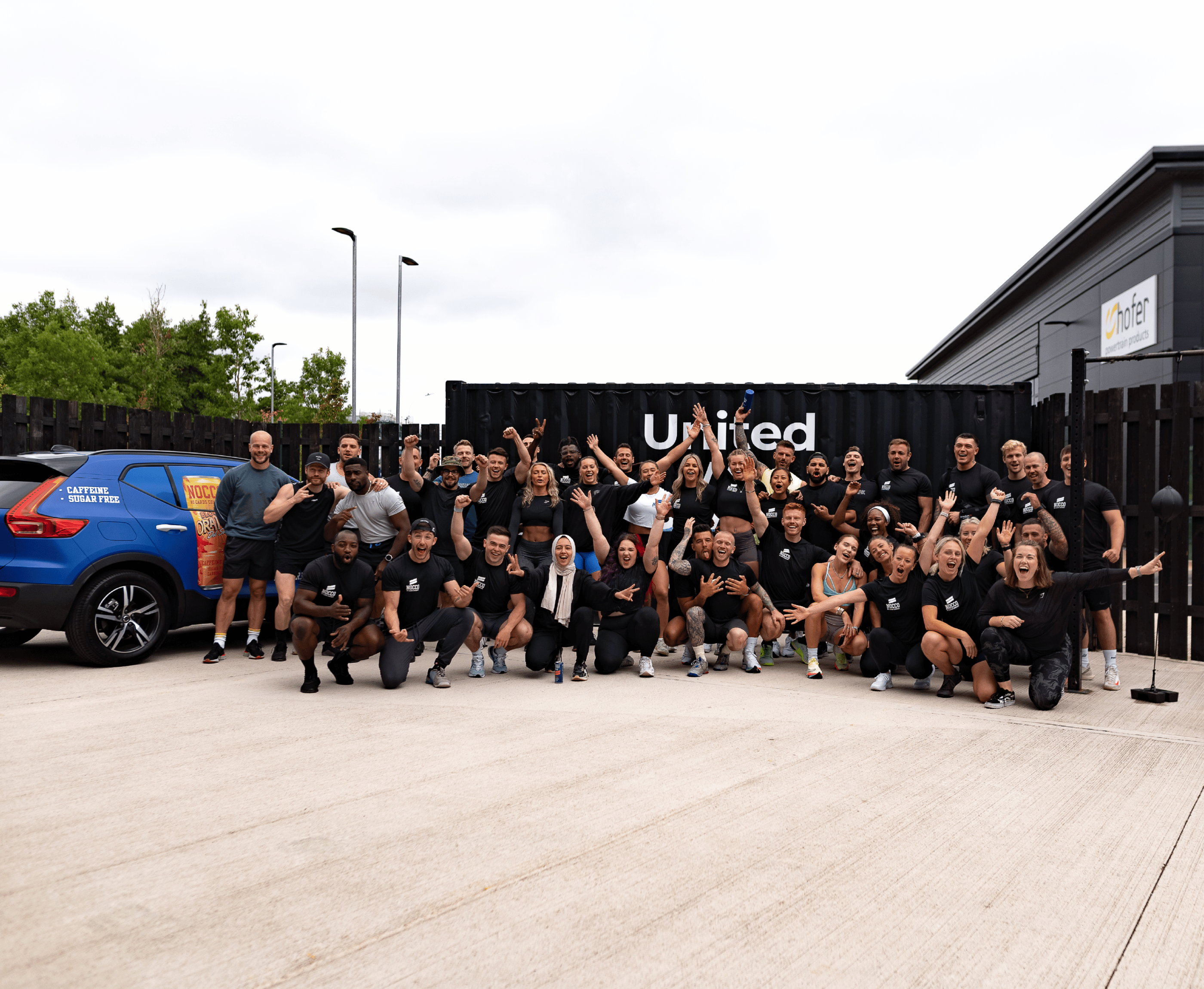 Group of joyful people posing outside a gym with a blue car on the left.
