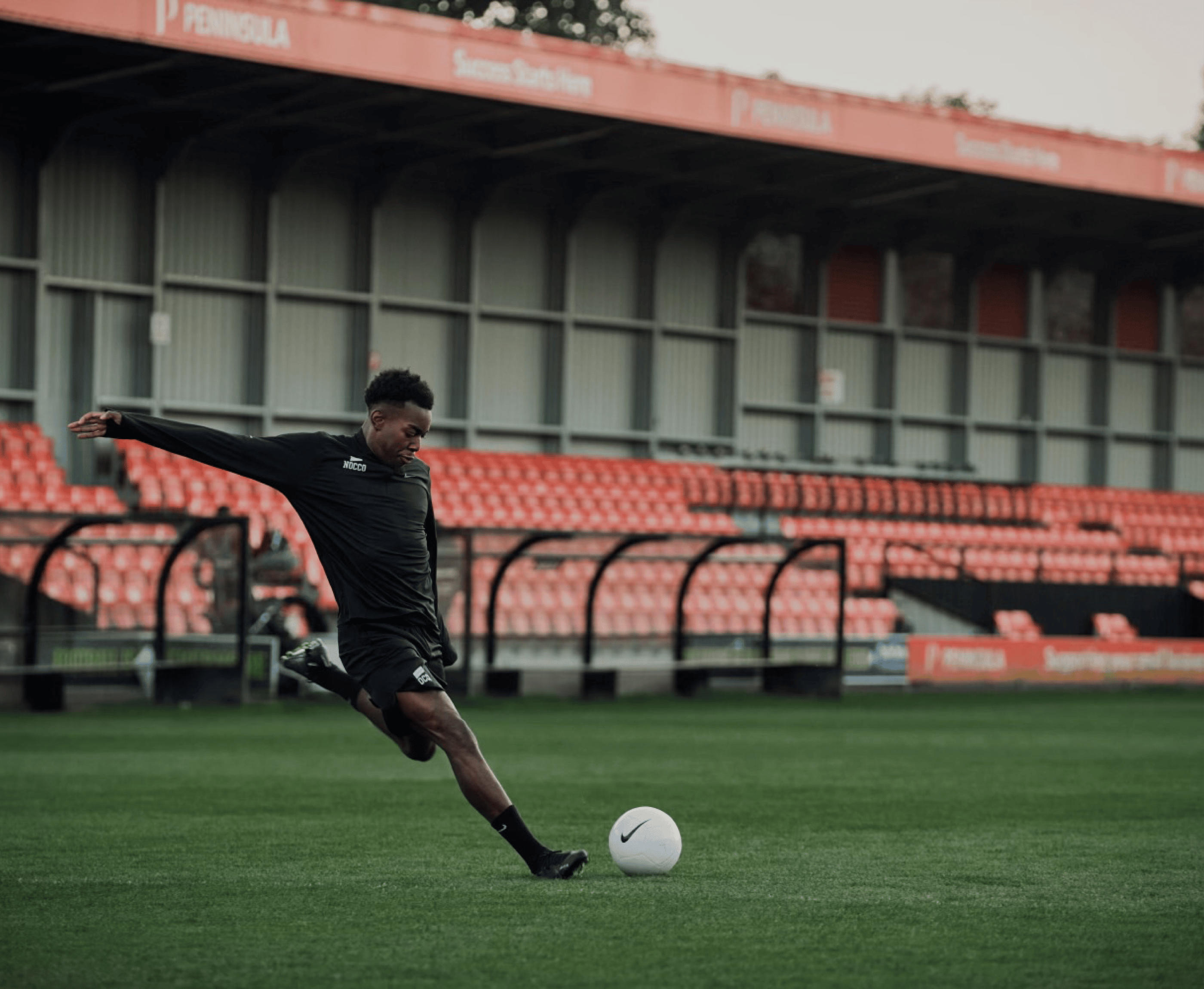 Soccer player kicking a ball on a field with empty red seats in background.